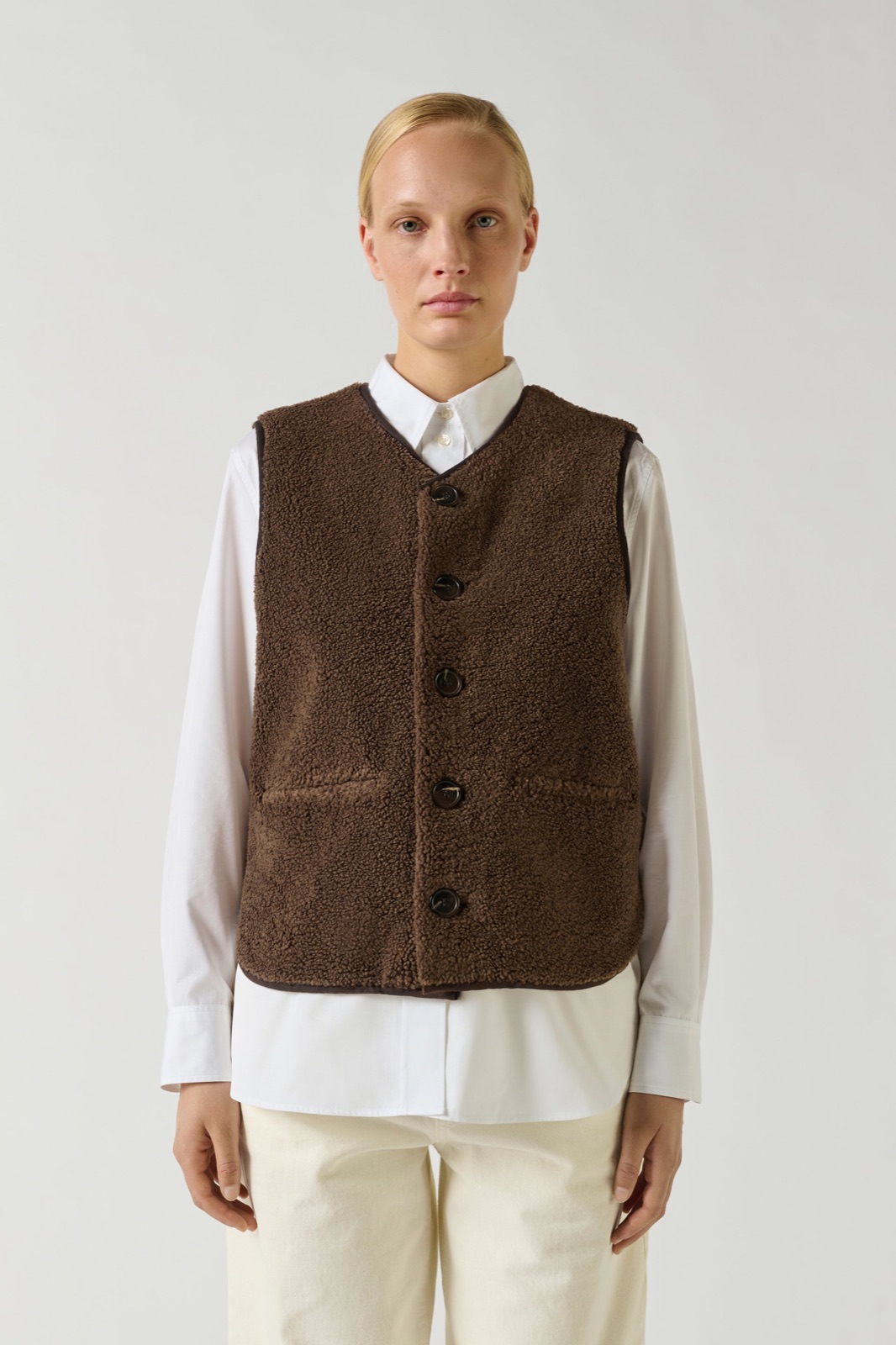 Gilet sans manches Fusil de Soeur en fausse fourrure brune, encolure en V et boutonnage devant, porté sur une chemise blanche