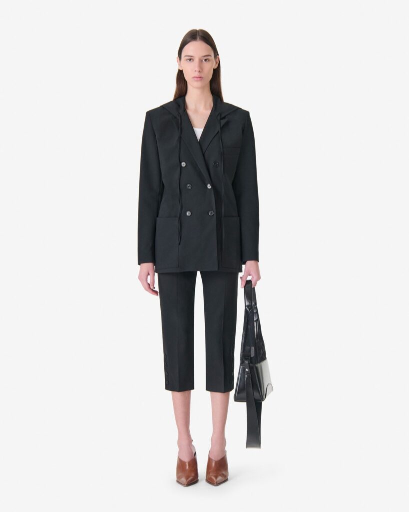 Veste de tailleur ALULA d'IRO en noir, coupe croisée avec capuche intégrée, portée sur un pantalon de tailleur noir