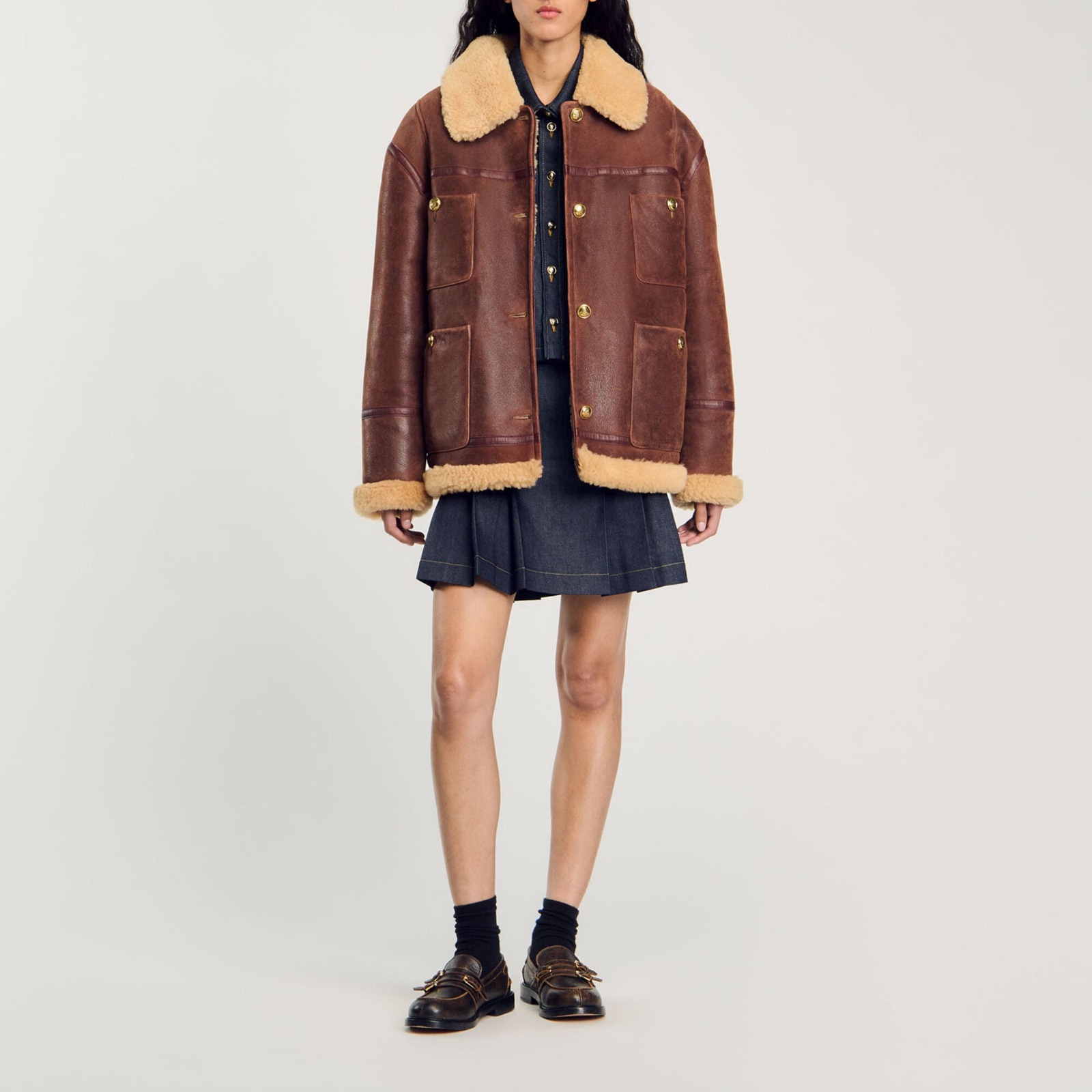 Veste en peau lainée marron Sandro, col et poignets en mouton crème, portée avec une jupe en jean plissée