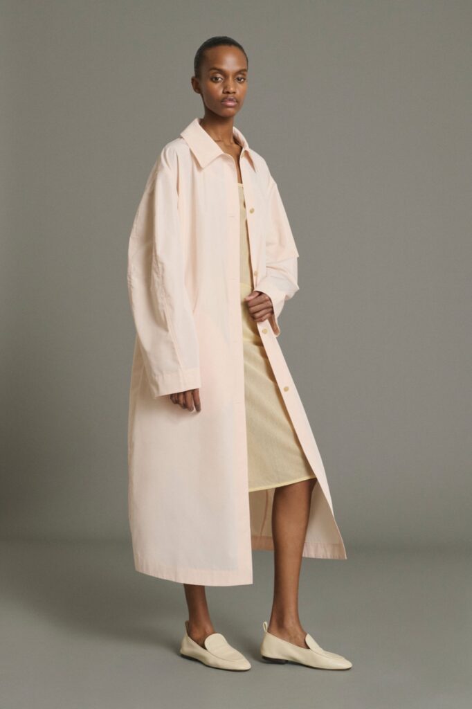 Manteau Joie de Soeur, long trench rose poudré porté ouvert sur une robe jaune pâle