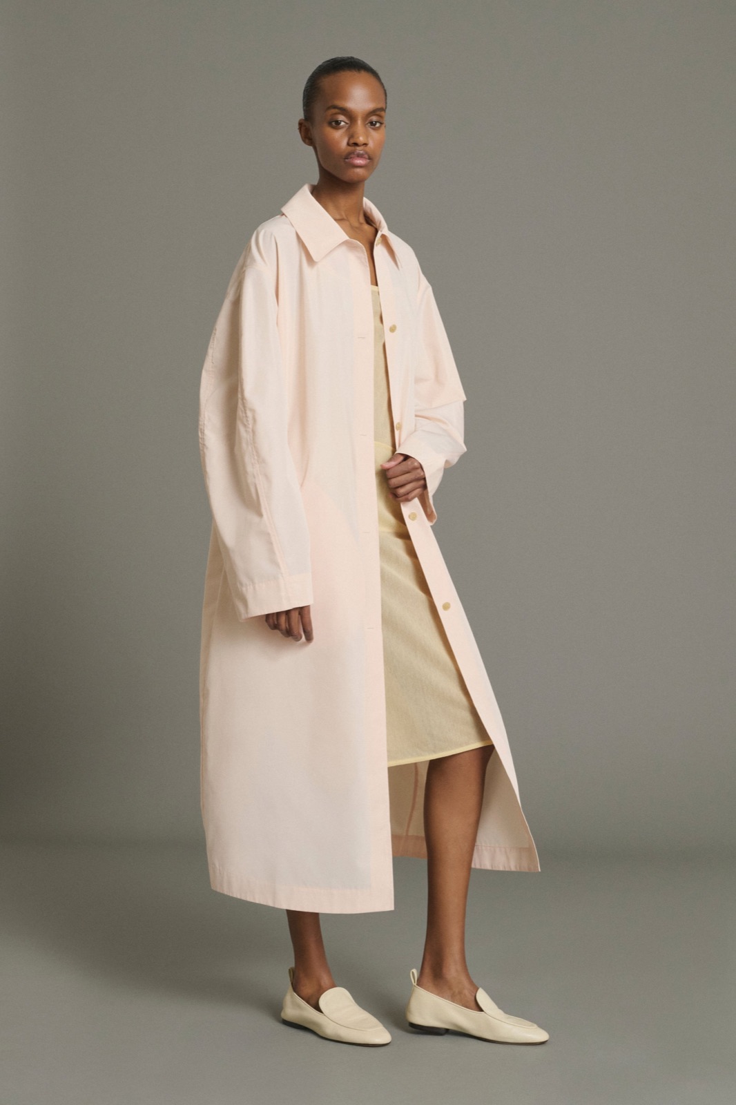 Manteau Joie de Soeur, long trench rose poudré porté ouvert sur une robe jaune pâle